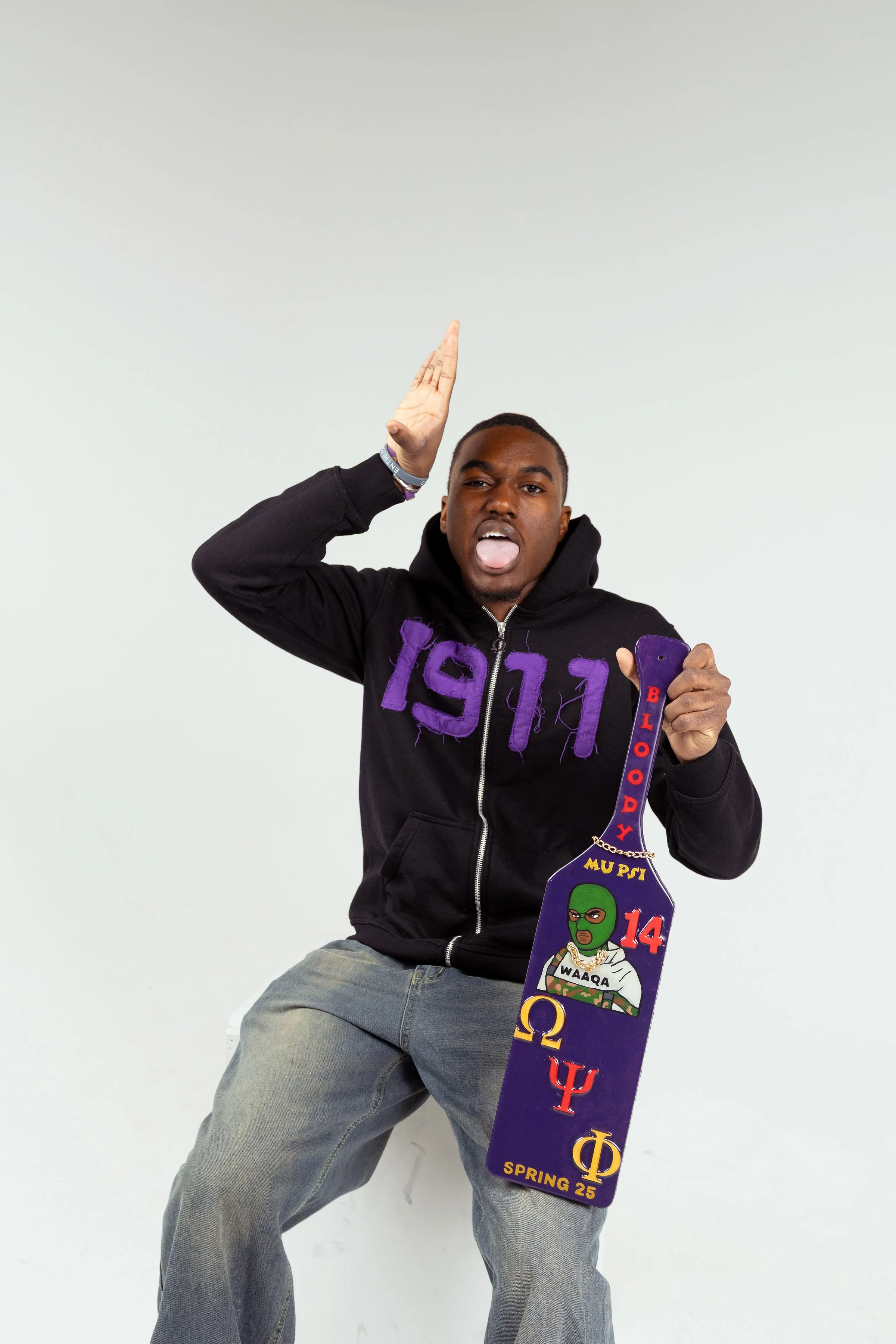 A young man with dark skin wearing a black hoodie with purple lettering, gray jeans, and various wristbands. He is sitting against a plain white background, holding a decorated skateboard with fraternity letters and a cartoon character, and making a playful gesture with his tongue out and one hand raised to his head.
