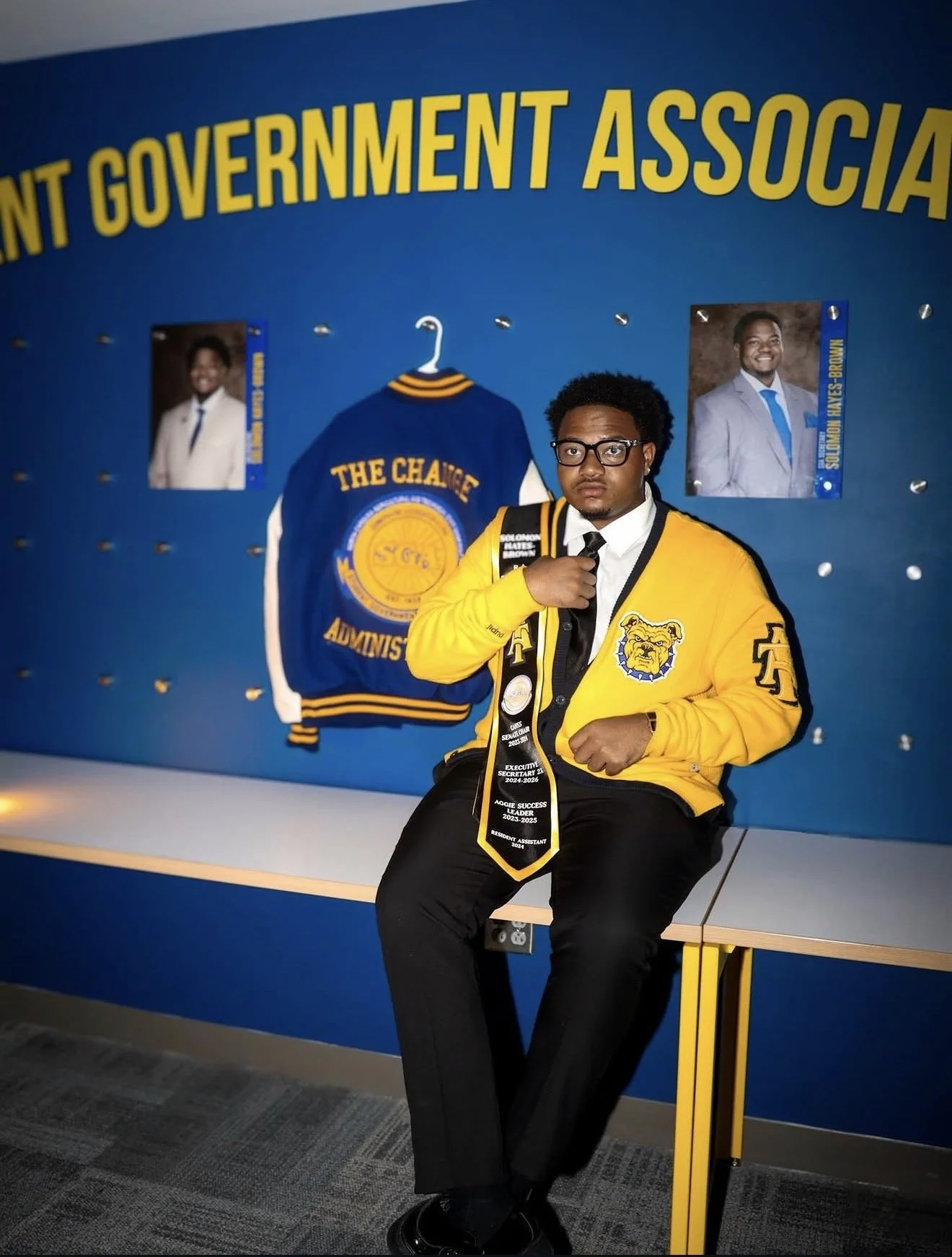 A young man with glasses wearing a yellow jacket and a necktie with various text, sitting on a table in front of a blue wall with pictures of two men and a varsity jacket. The wall features large yellow text reading 'STUDENT GOVERNMENT ASSOCIATION'.