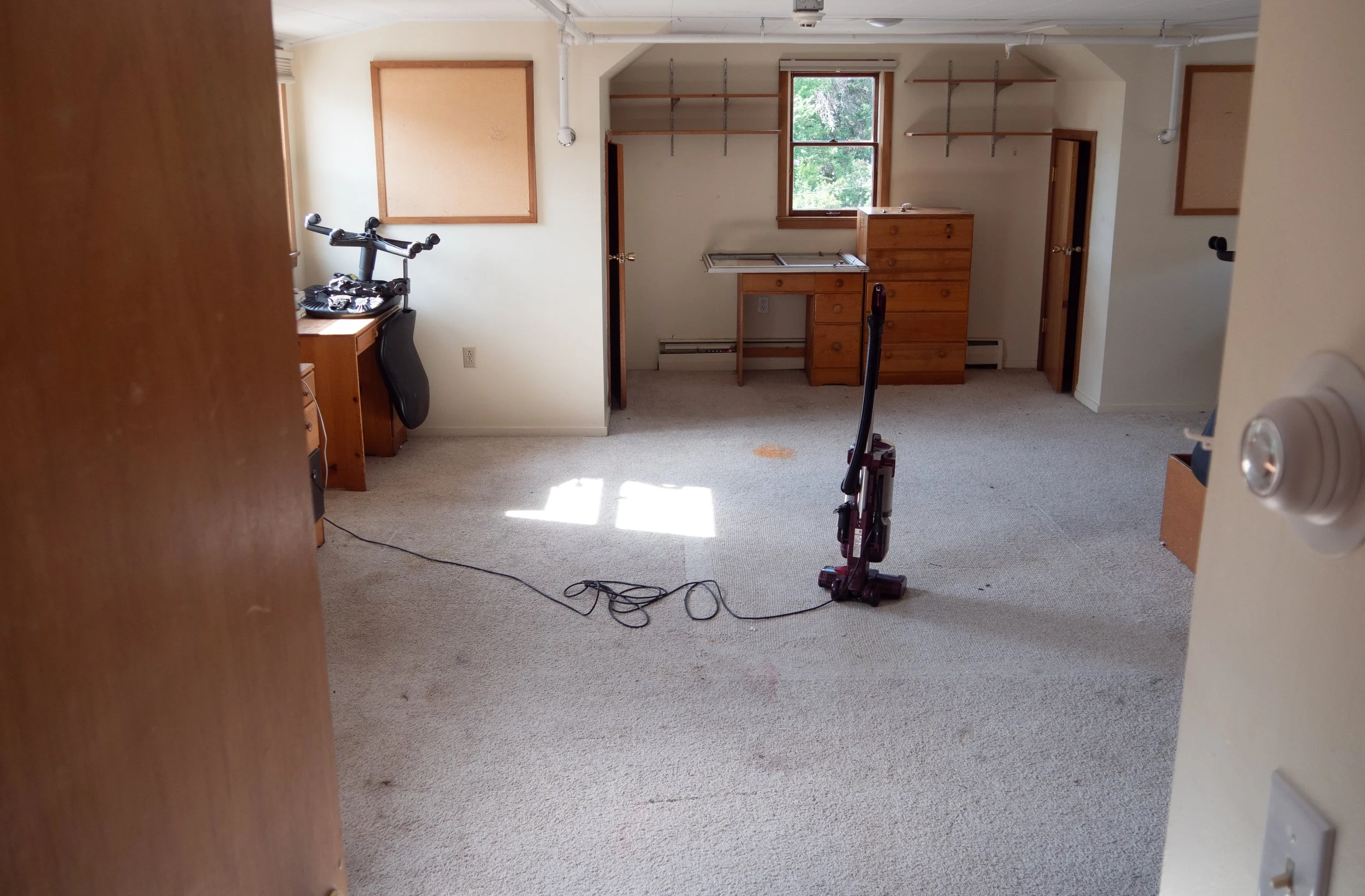 Empty room with beige carpet, two wooden dressers, ceiling shelves, a window with green trees outside, a vacuum cleaner, and some scattered cables. Eco-friendly carpet cleaning solutions safe for pets and children in Belgrade.