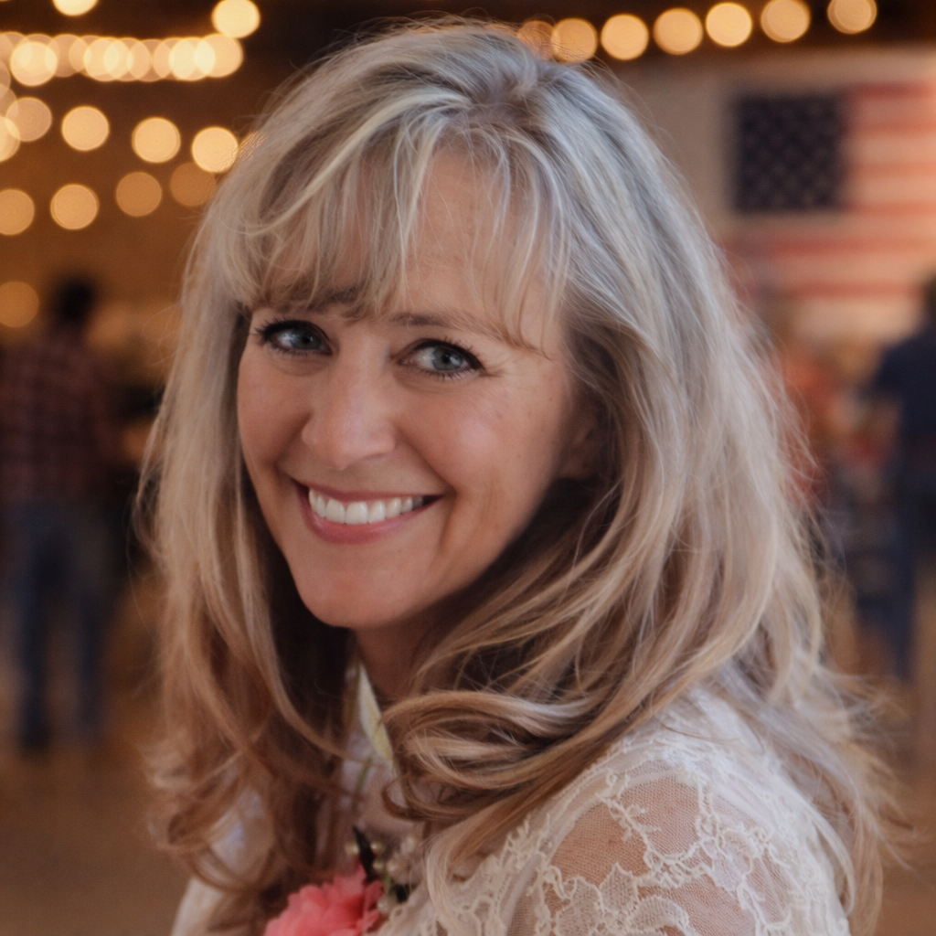 A smiling woman with blonde, wavy hair in a well-lit indoor setting with string lights in the background.