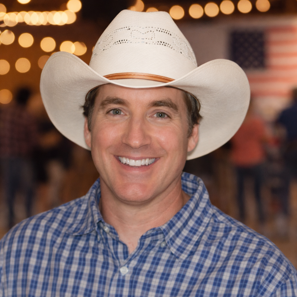A man smiling, wearing a white cowboy hat and a blue checkered shirt, at a festive event with blurred background lights and American flag decorations.