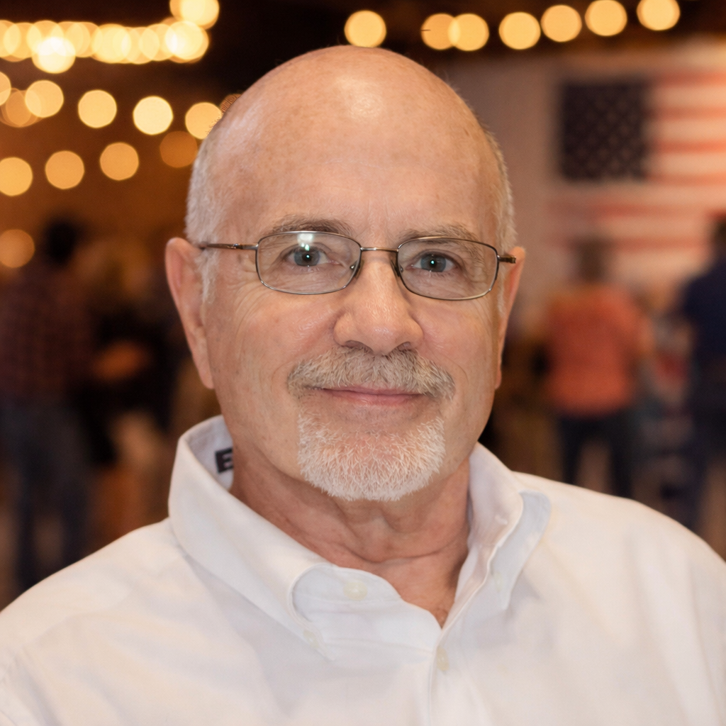 Close-up of an older man with glasses, a white shirt, and a light goatee, smiling at a social event with blurred string lights and American flags in the background.