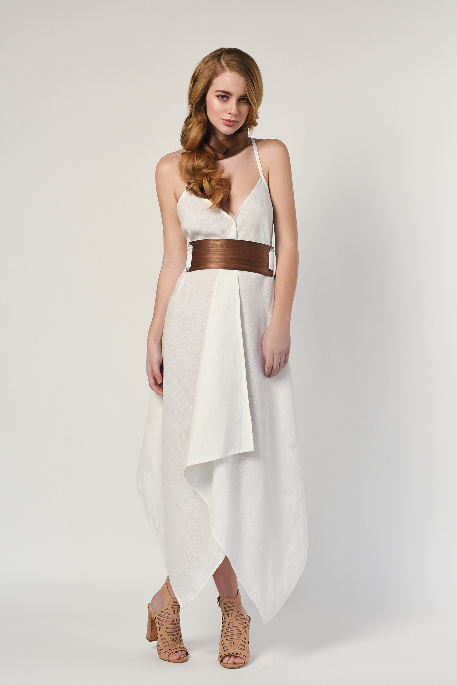 A woman with red hair styled in loose curls, wearing a white sleeveless dress with thin straps, a brown wide belt, and tan open-toe high-heeled shoes, standing against a plain white background.