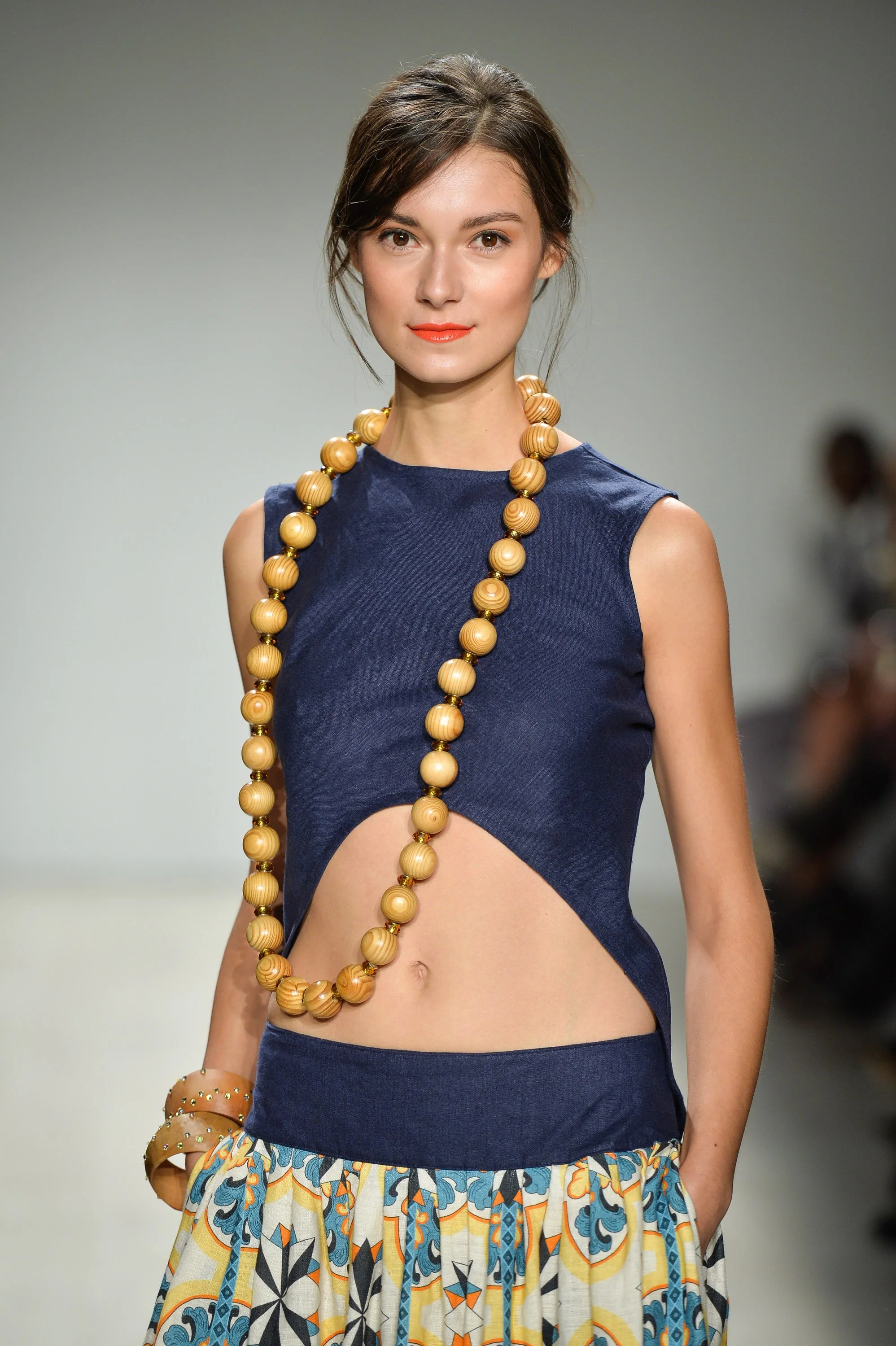 A woman walking on the runway during a fashion show, wearing a sleeveless blue crop top with a cutout at the midriff, a long beaded necklace, and a colorful patterned skirt.