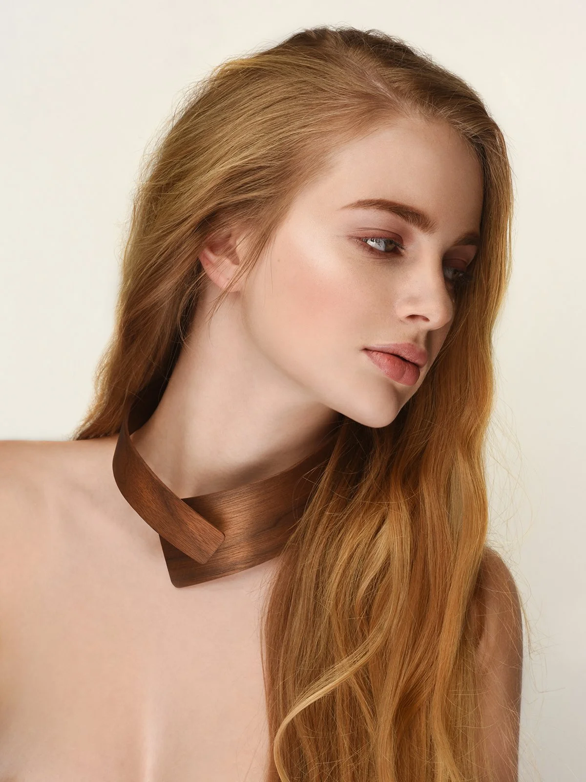 A young woman with long, wavy red hair looking down, wearing a brown ribbon around her neck, against a neutral background.