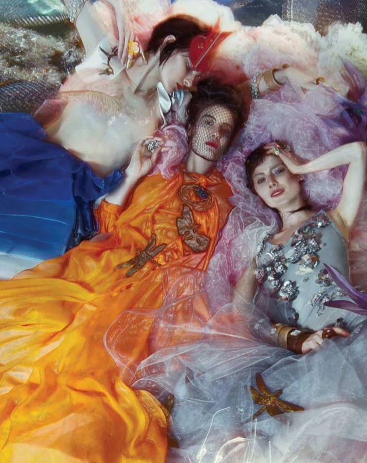 Three women in colorful, elaborate dresses lying on the ground, with their heads together. They are surrounded by sheer fabric and decorated with butterflies and accessories.