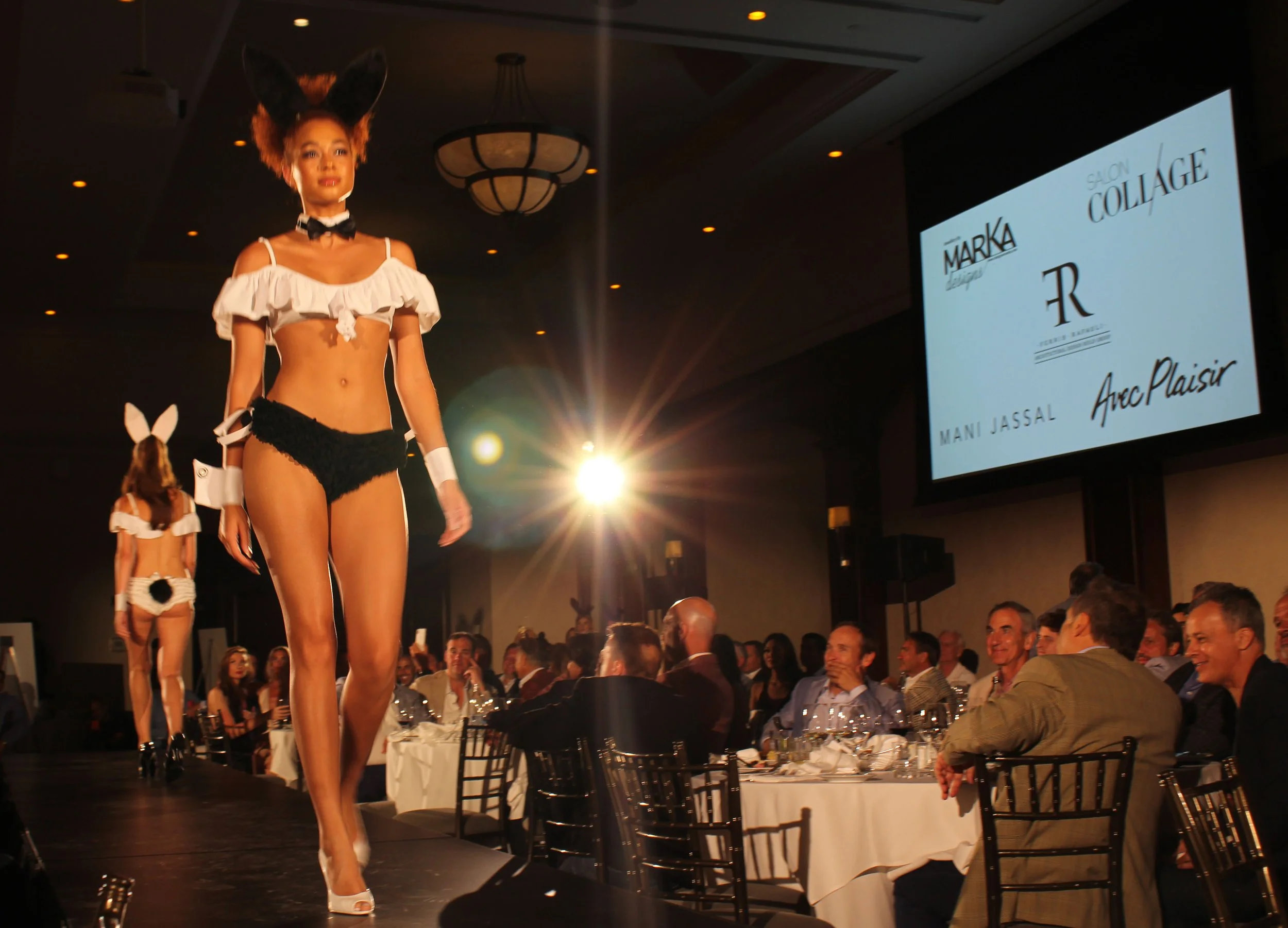 Models walking on runway during a fashion show, with audience seated at tables in the background, and a large screen displaying brand logos and designer names.