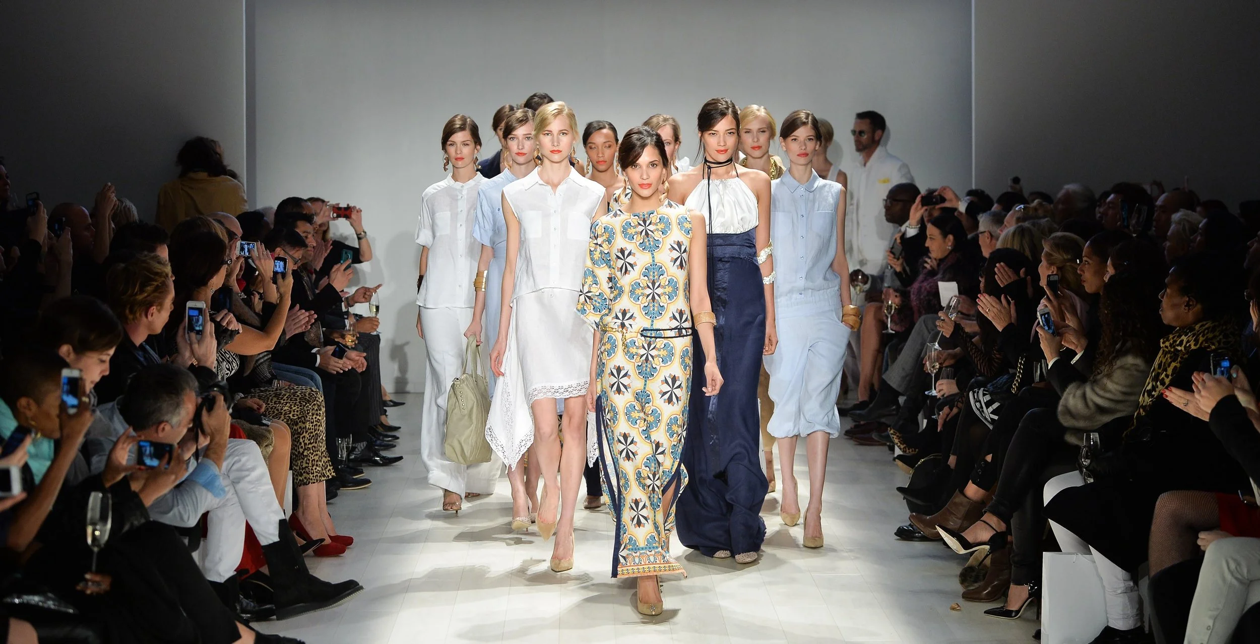 Models walk down the runway at a fashion show, with audience members seated on both sides capturing the event on their phones.