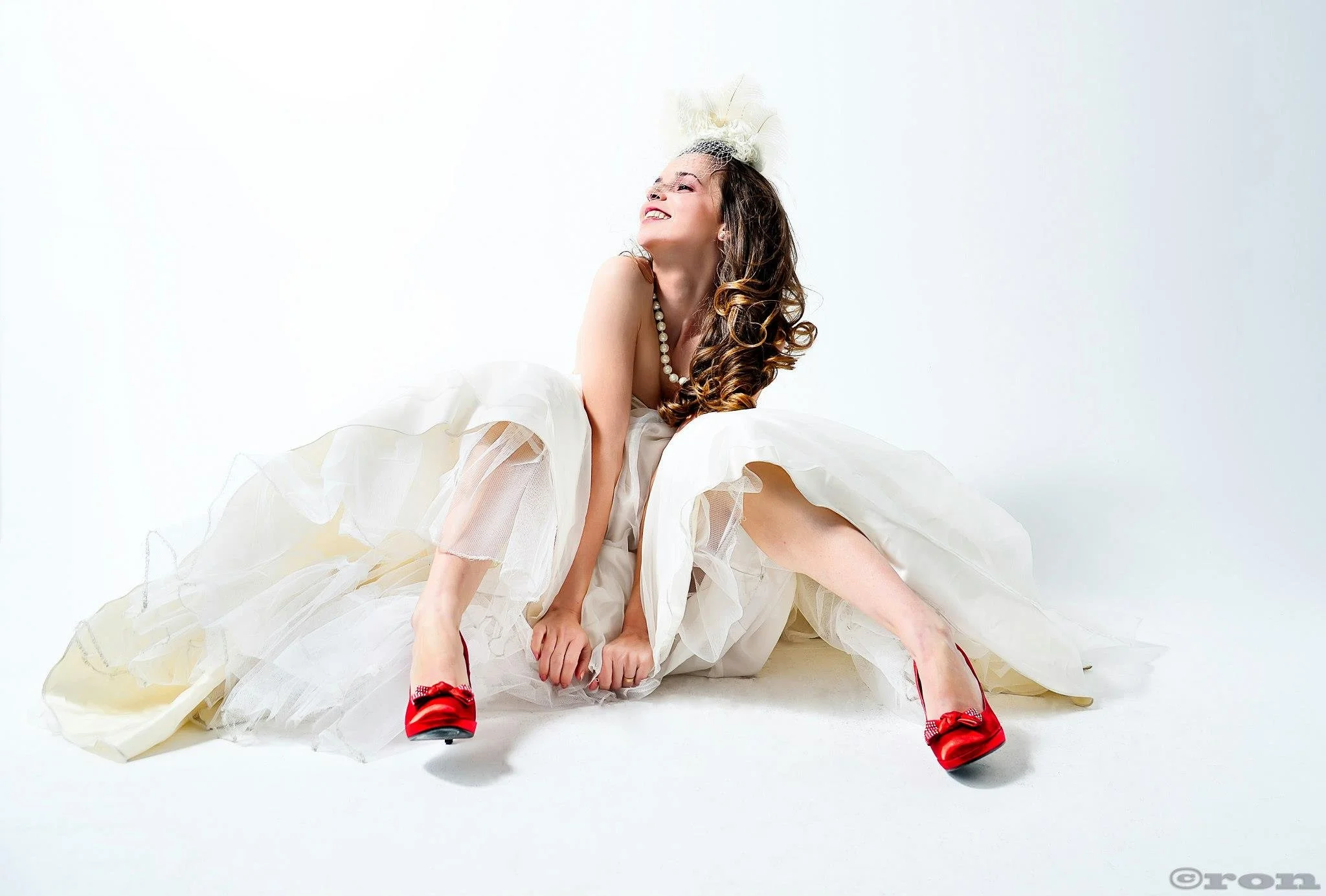 Woman in a white wedding dress with red shoes, sitting on the floor with legs apart, smiling, with curly hair, wearing a pearl necklace and a feathered headpiece.