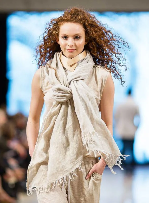 A young woman with curly red hair walking on a runway during a fashion show, wearing a beige sleeveless top, loose beige pants, and a large, light-colored scarf draped around her neck and shoulders.
