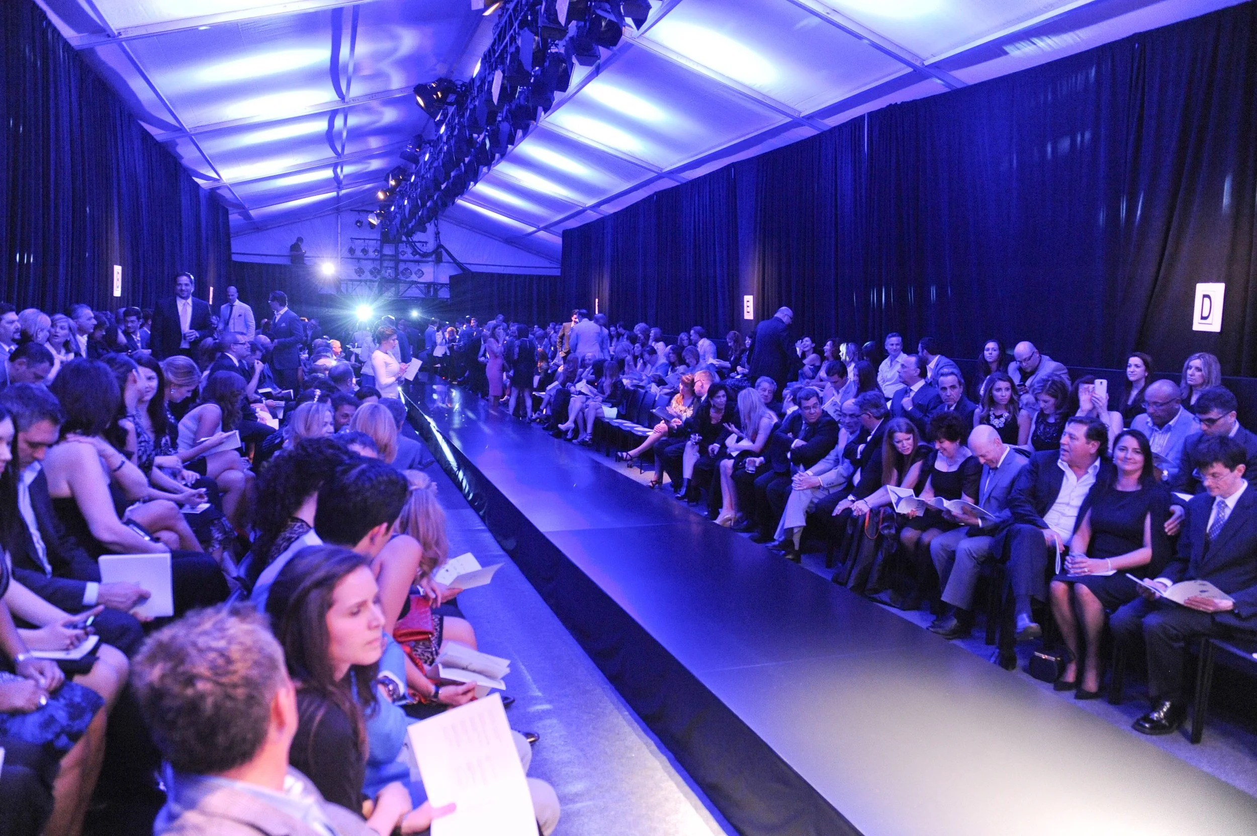 People attending a fashion show seated along a runway inside a large tent with blue lighting.