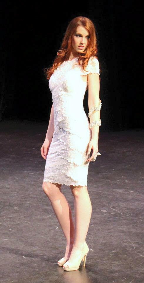A woman with red hair wearing a white lace dress and high heels on a dark stage.
