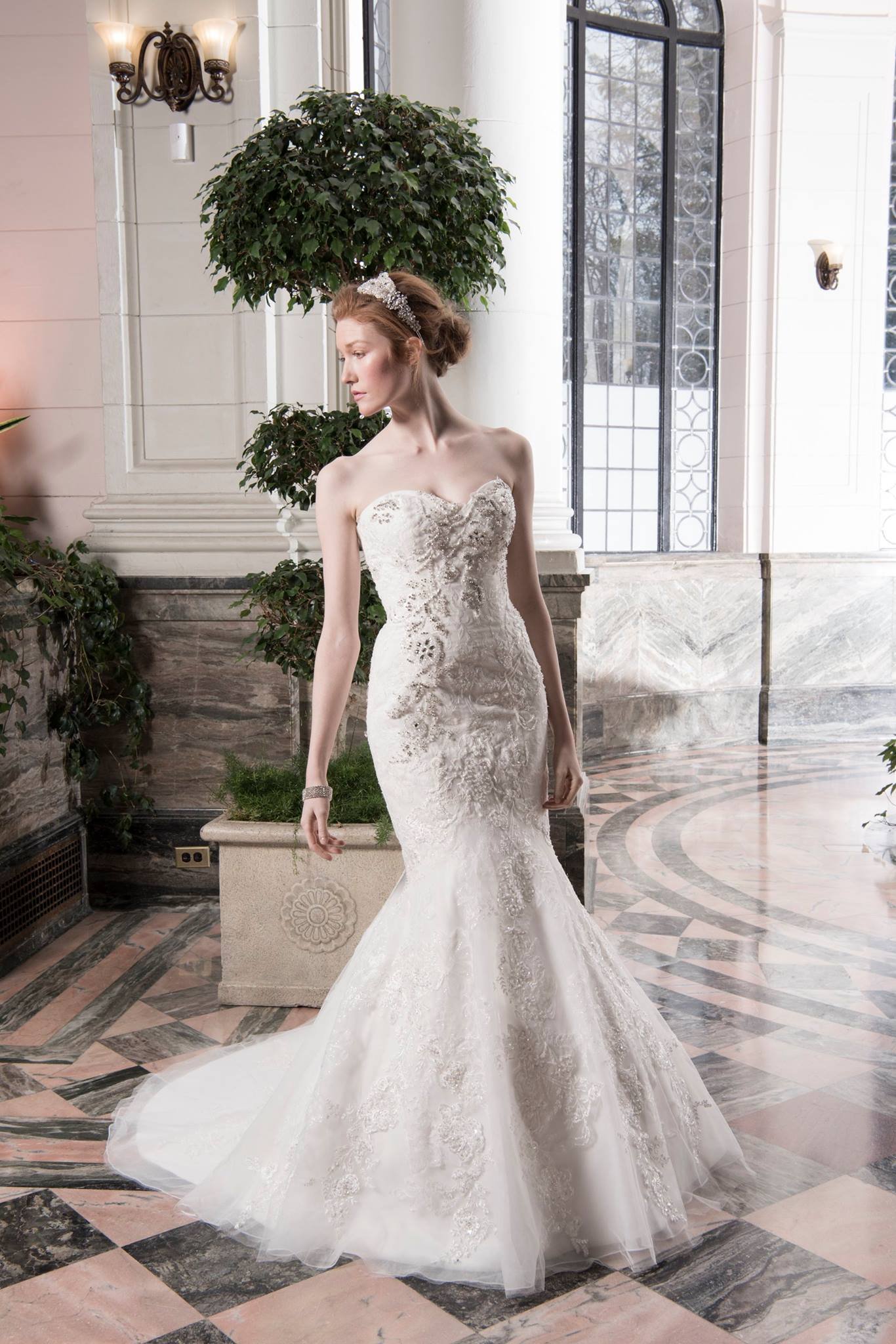 A woman wearing a white wedding dress with lace details, standing indoors near a window and potted plants.