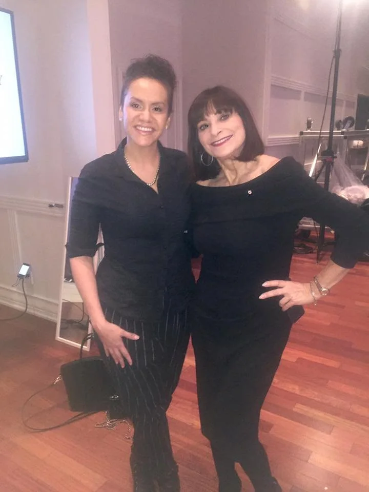 Two women standing close together, smiling, in a room with wooden floors and a TV screen to the left.