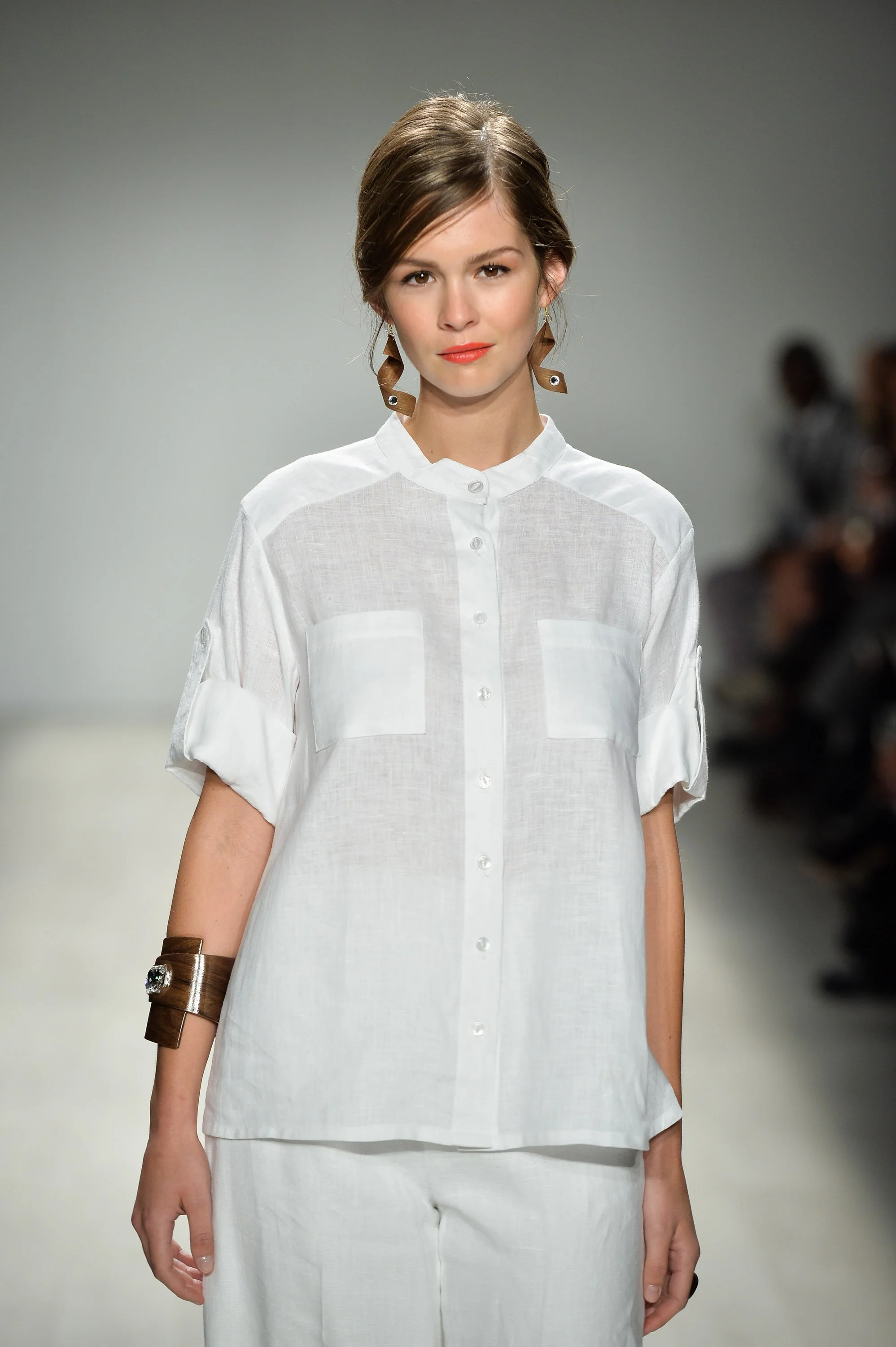 Model walking on runway in white linen shirt with rolled sleeves and matching white pants, wearing brown earring and bracelet, during a fashion show.