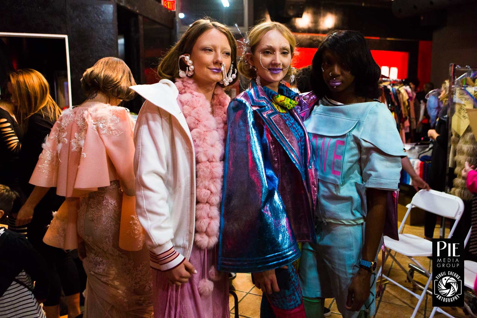 Four women standing together at a fashion event, wearing colorful and avant-garde outfits with bold accessories, in an indoor setting with racks of clothing and other people in the background.