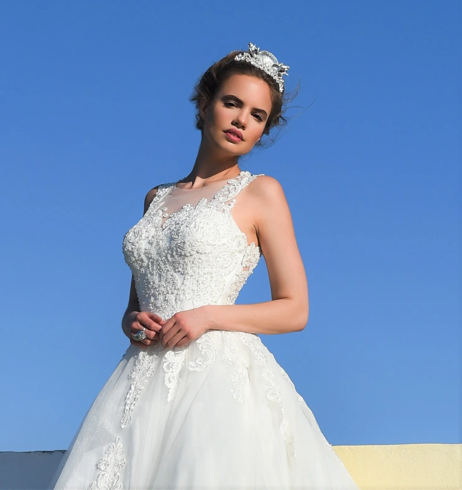 A woman in a detailed white wedding dress with lace and embroidery, wearing a tiara, stands against a bright blue sky.