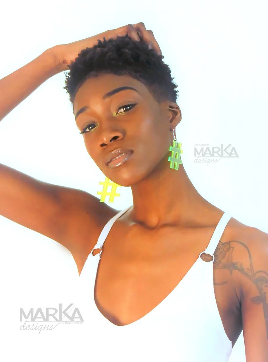 A woman with short curly hair wearing large neon green hashtag earrings and a white top, posing against a white background.