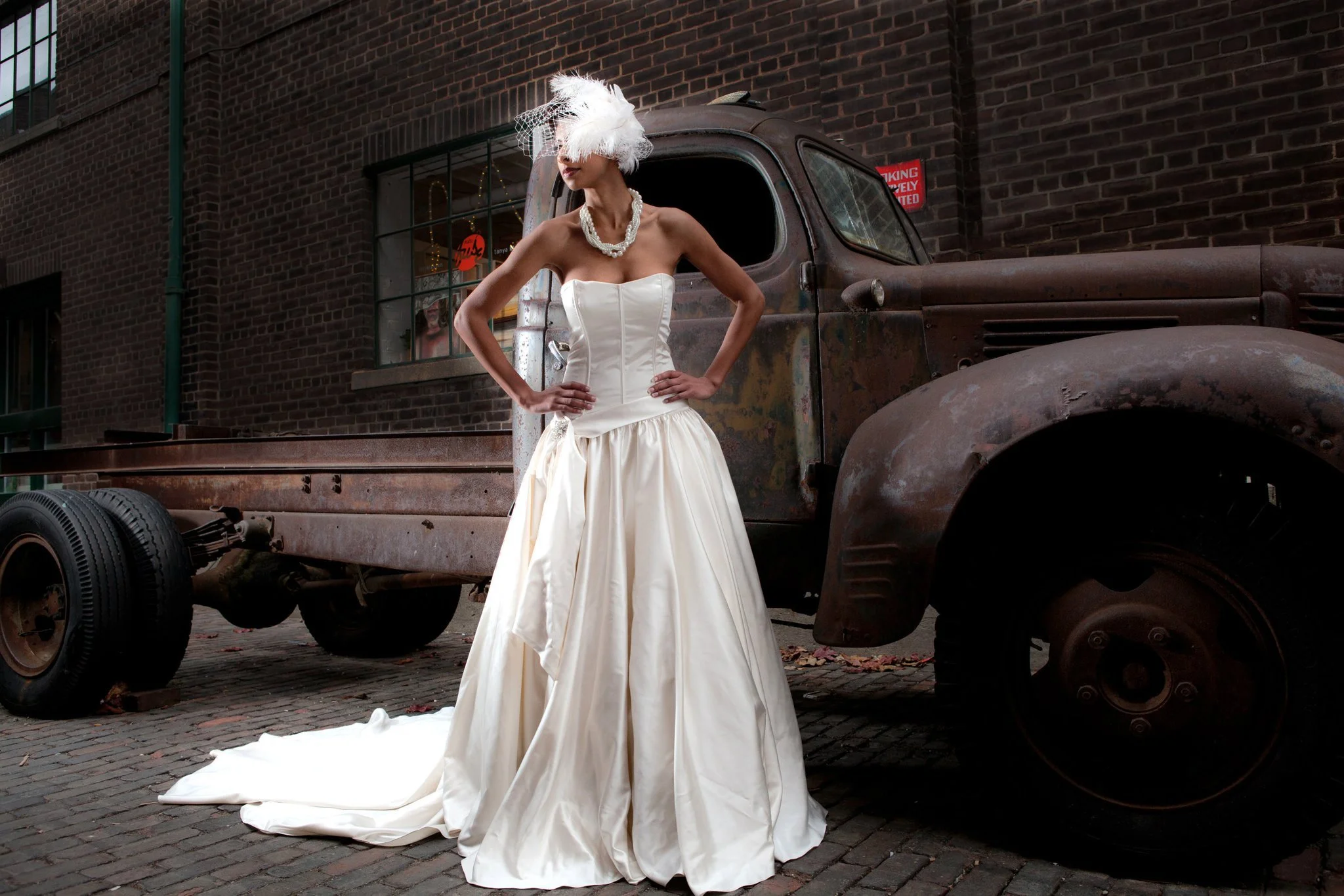 A woman in a white wedding gown, veil, and feathered headpiece standing next to an old rusty truck on a cobblestone street.