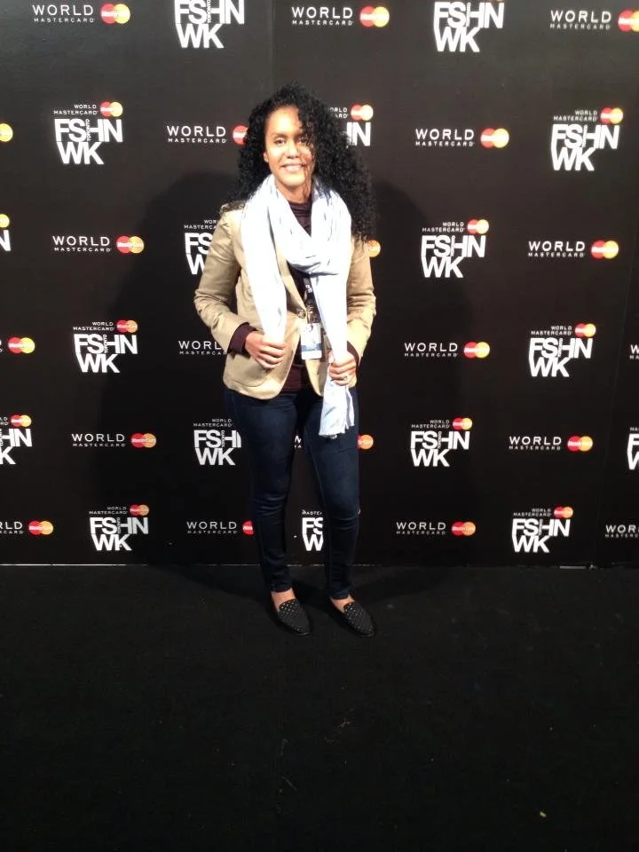 A woman standing in front of a black step-and-repeat backdrop with logos for FASHWK, WORLD MASTERCARD, and Mastercard, at an event. She has curly black hair, is smiling, and is wearing a beige blazer, dark jeans, black shoes with embellishments, and 
