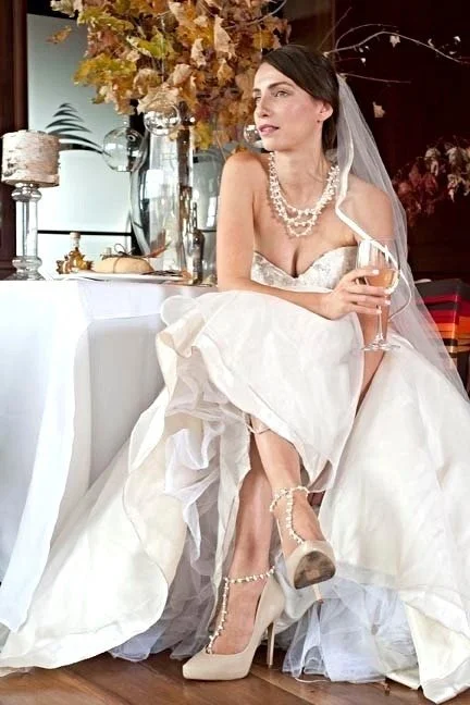 Bride in wedding dress sitting on a white chair, holding a glass of champagne. Decorated with pearls, surrounded by elegant decorations and autumn-themed leaves.