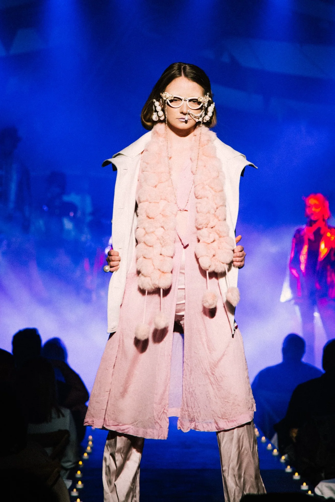 A fashion model walking on runway wearing pink layered outfit with fluffy scarf and statement glasses, with audience and blue background.