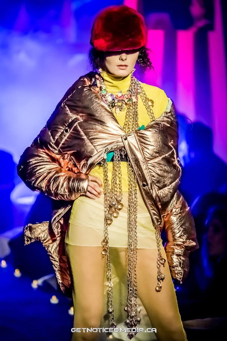 Fashion model walking on runway wearing gold metallic jacket, yellow top with colorful jewelry, and a red fur hat in a vibrant, colorful setting.