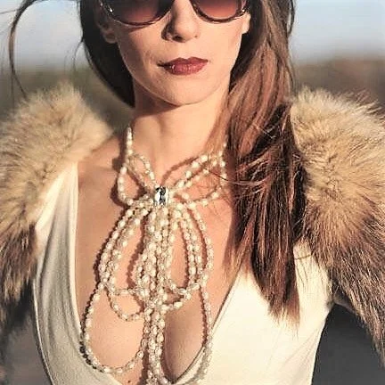 A woman wearing sunglasses, a white top, a fur coat, and multiple pearl necklaces outdoors.