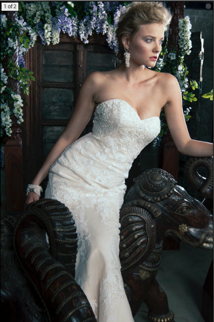 A woman in a strapless white wedding gown with lace details poses indoors, surrounded by floral arrangements and dark wooden furniture.