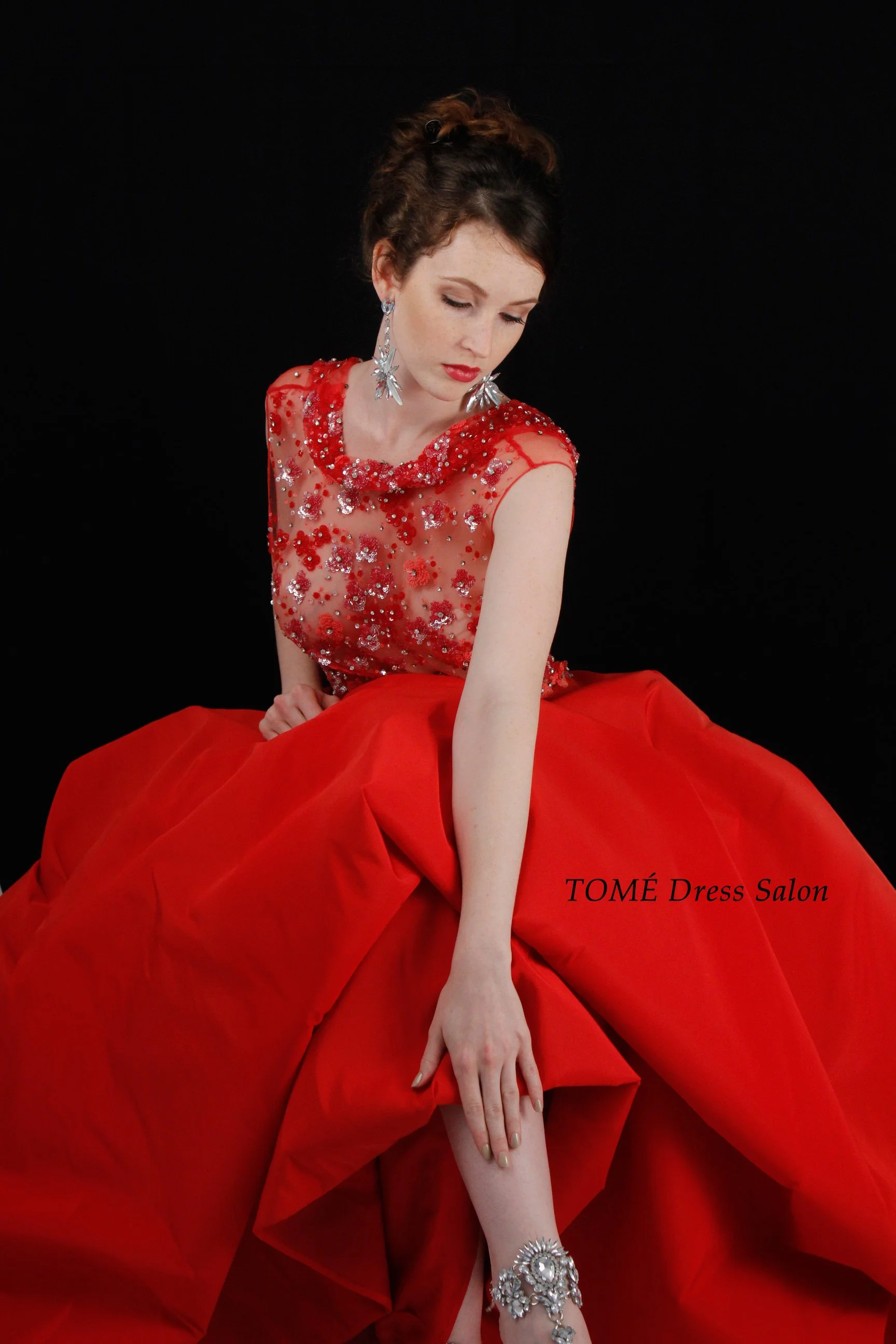 A young woman poses wearing an elegant red gown with sparkly embellishments on the top and a voluminous skirt, against a black background. She has styled hair, large star-shaped earrings, and a decorative bracelet.