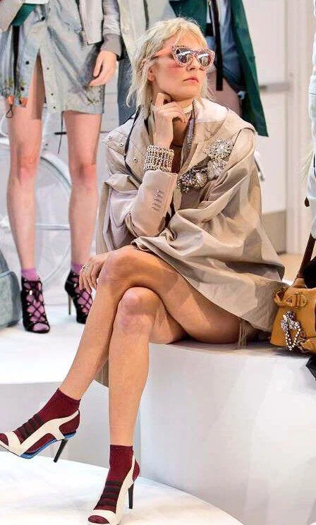 Fashion model sitting on a white stage at a fashion show, wearing a beige oversized coat with embellishments, red socks, and high heels, with a woman standing and other models in the background.