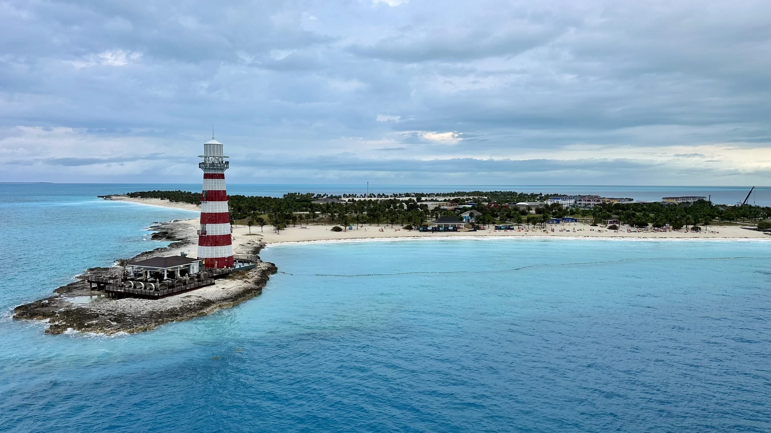 A lighthouse with red and white stripes on a small rocky island off the coast of a sandy beach with clear blue water. The background shows a beach with palm trees, buildings, and a cloudy sky. Michael and Tiffany Travels MSC Ocean Cay Seashore MSC