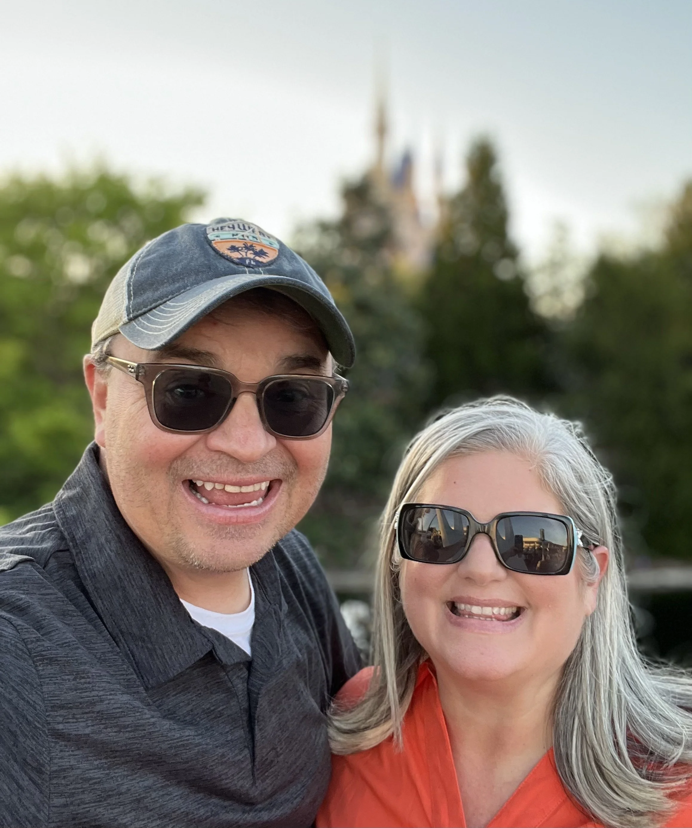 A smiling man and woman wearing sunglasses taking a selfie outdoors with a blurred castle and trees in the background. Michael and Tiffany Travels Magic Kingdom Walt Disney World Cinderella’s Castle
