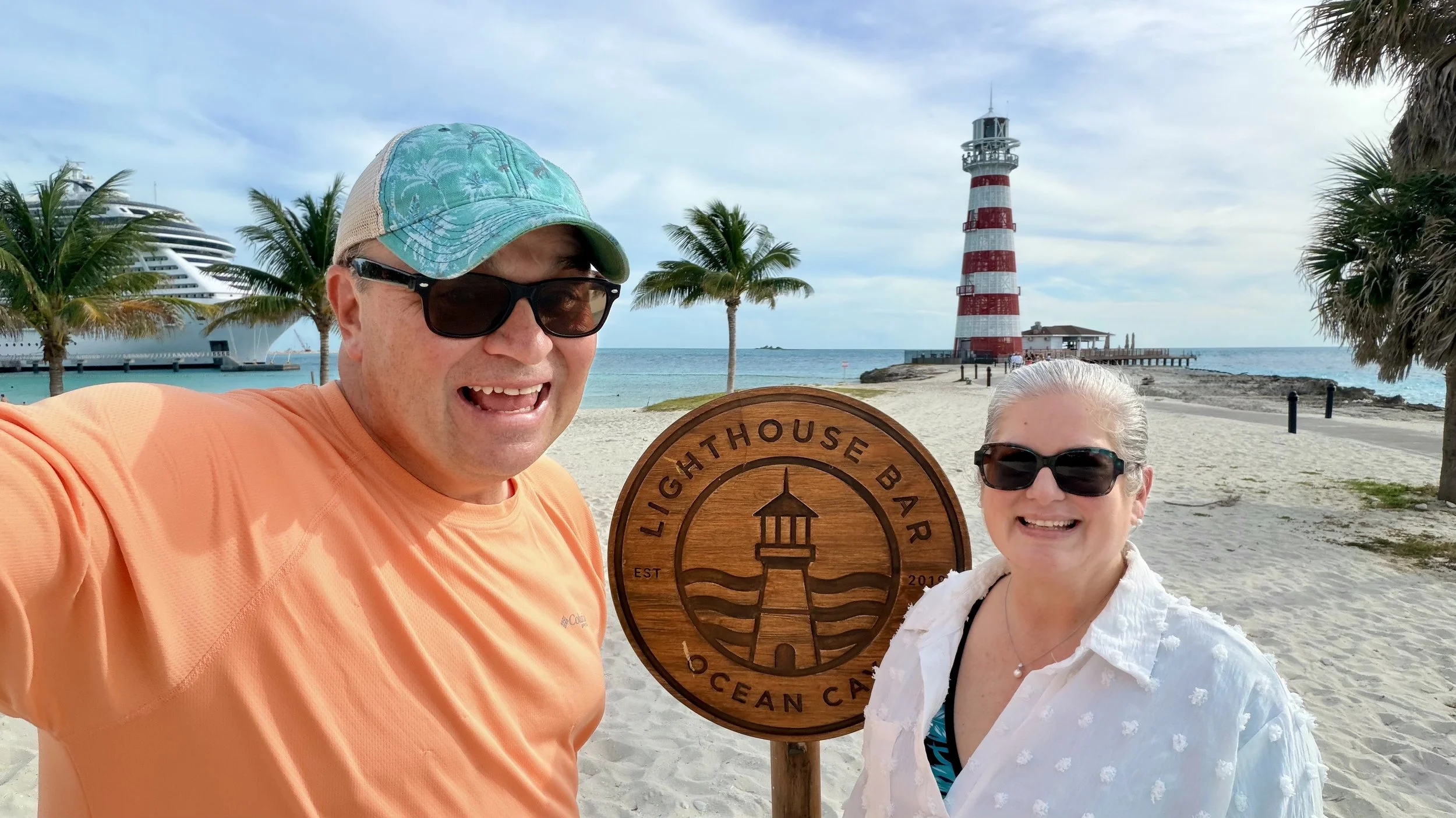 Two smiling people wearing sunglasses on a sandy beach near a lighthouse, holding a wooden sign for Lighthouse Bar Ocean Café, with a cruise ship and palm trees in the background. Michael and Tiffany Travels MSC Cruises Ocean Cay Lighthouse Beach.