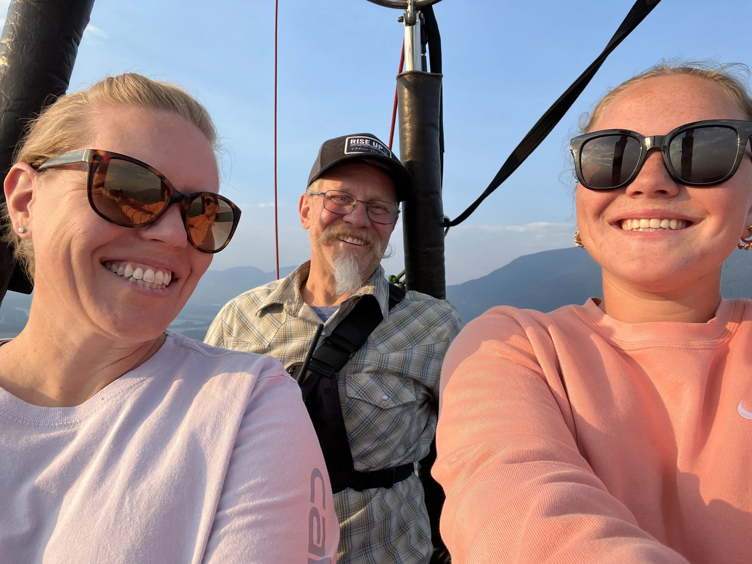 Three smiling people taking a selfie, with two women wearing sunglasses and a man with a beard and glasses in the middle, against a backdrop of mountains and a blue sky.