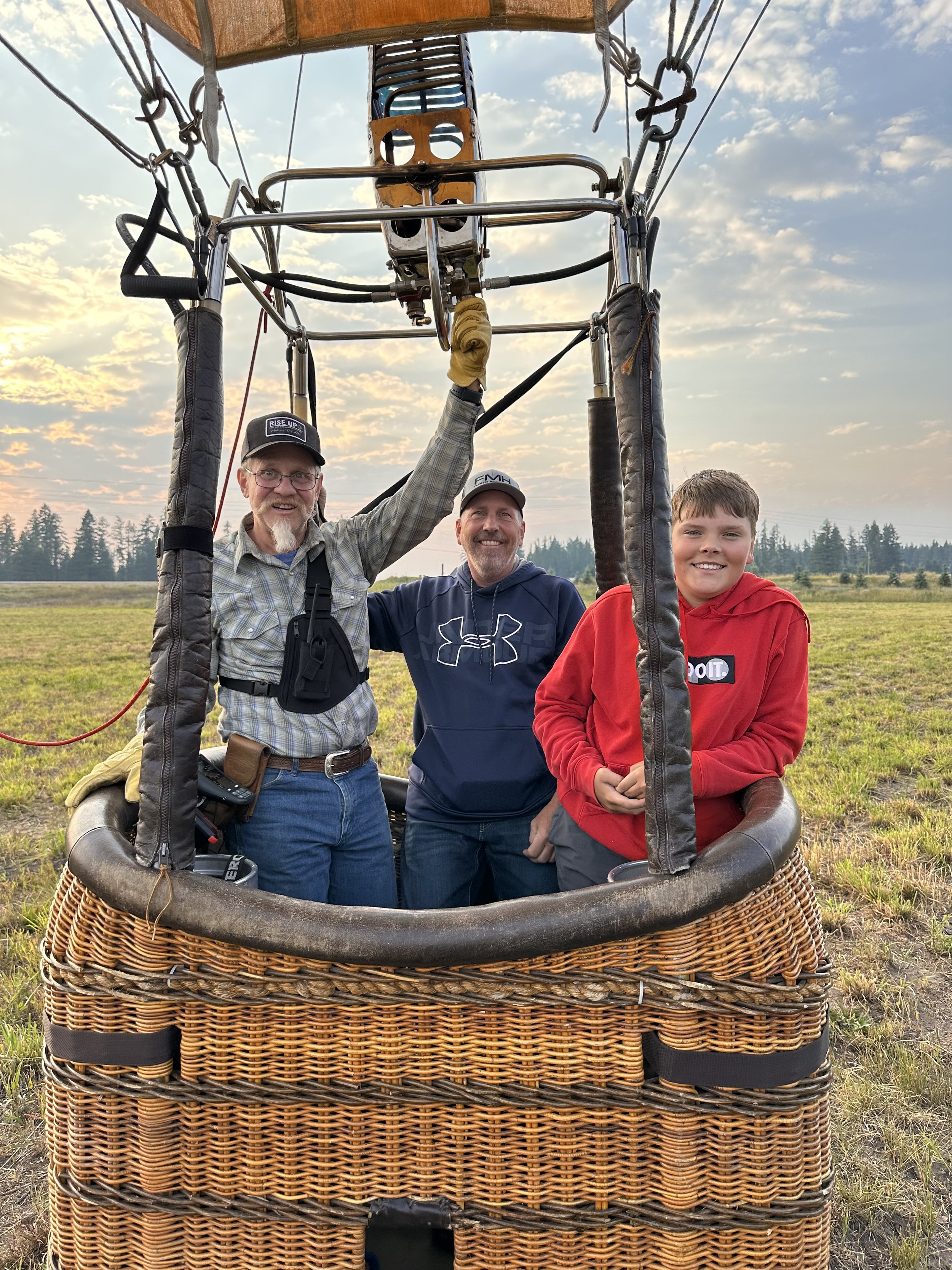 Three people standing inside a hot air balloon basket in an open field with a blue sky and scattered clouds. The basket is woven wicker with a metal frame, and the hot air balloon's envelope is partially visible above them.