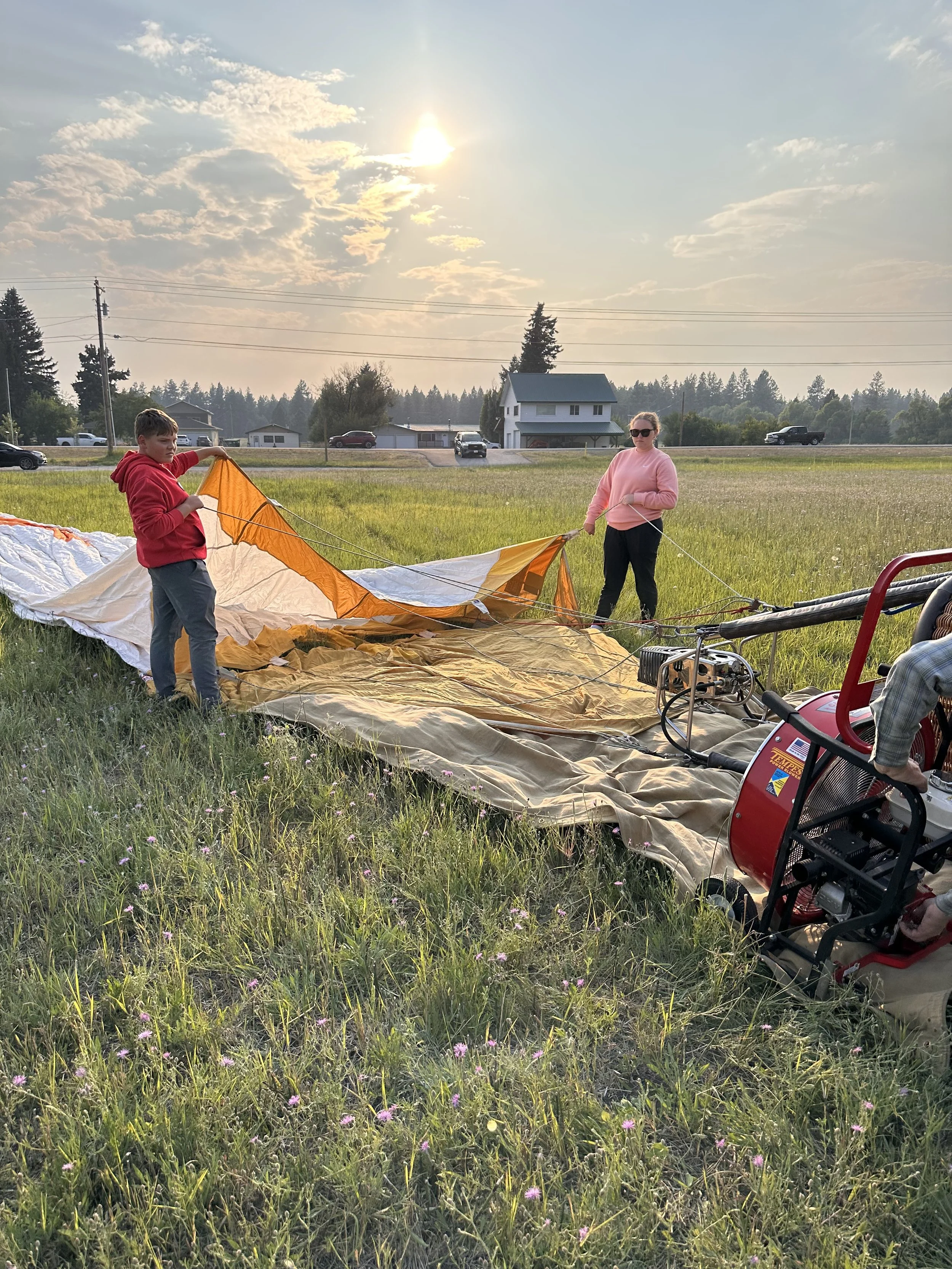 Two people preparing a hot air balloon for takeoff in a grassy field during sunset, with houses and trees in the background.