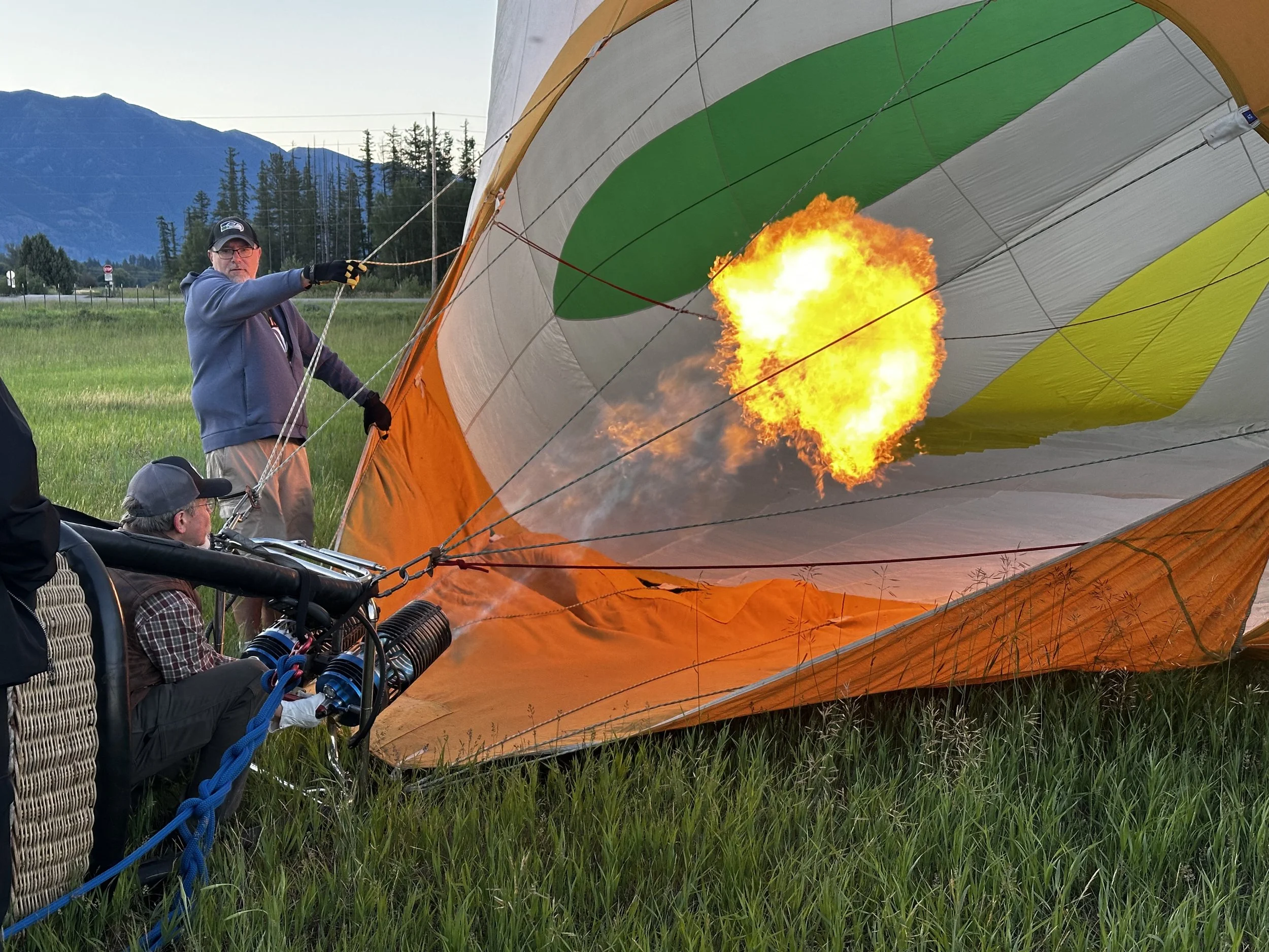 A hot air balloon is inflating on a grassy field with a mountain and trees in the background. People are working on the balloon, with one man holding the fabric and the pilot operating the burners, which are producing flames.