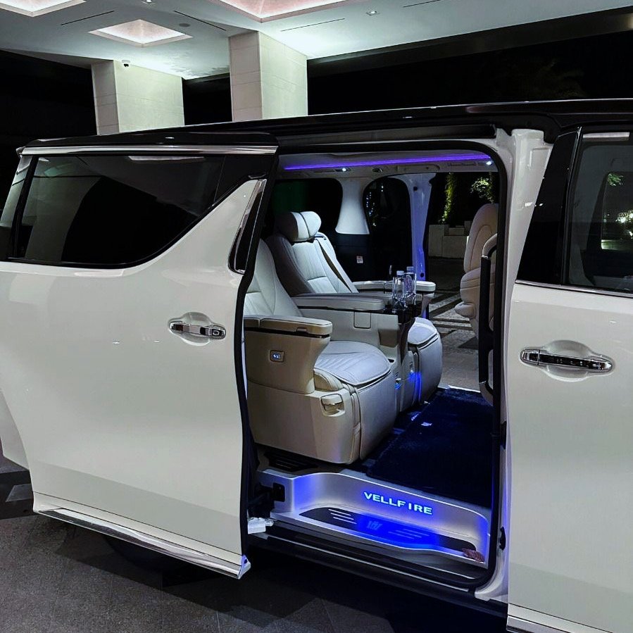 Luxury white van with open sliding door showing interior with beige leather seats, built-in controls, and blue ambient lighting at night.