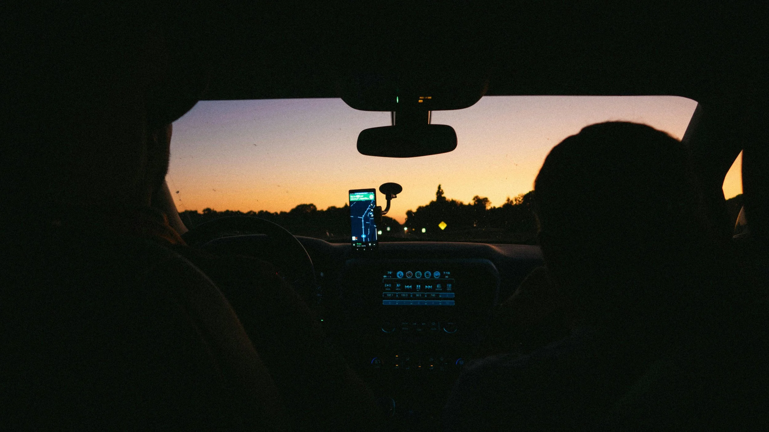 Interior of a car at dusk with two passengers, dashboard illuminated, GPS screen visible, and orange and purple sunset sky outside.