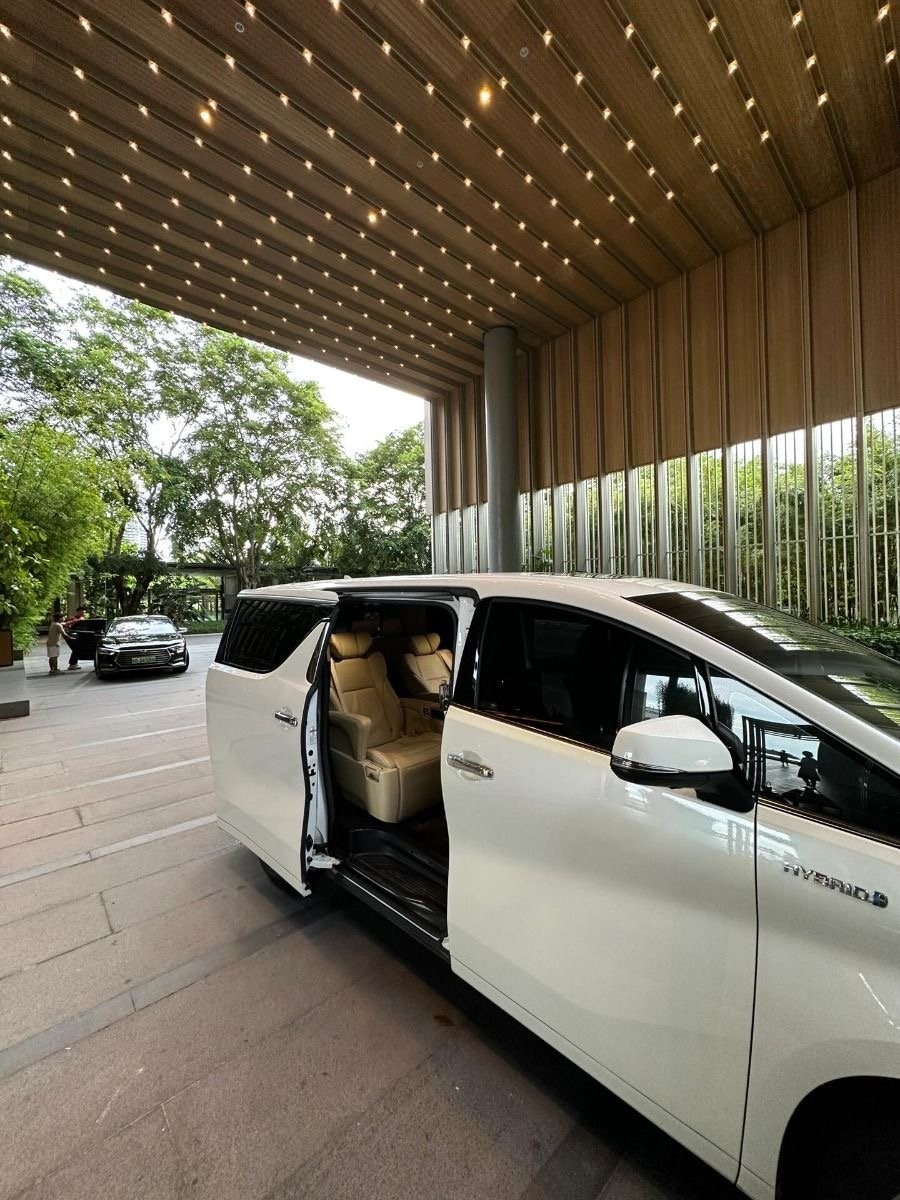 White hybrid minivan with beige interior parked under a modern, illuminated canopy with trees in the background.