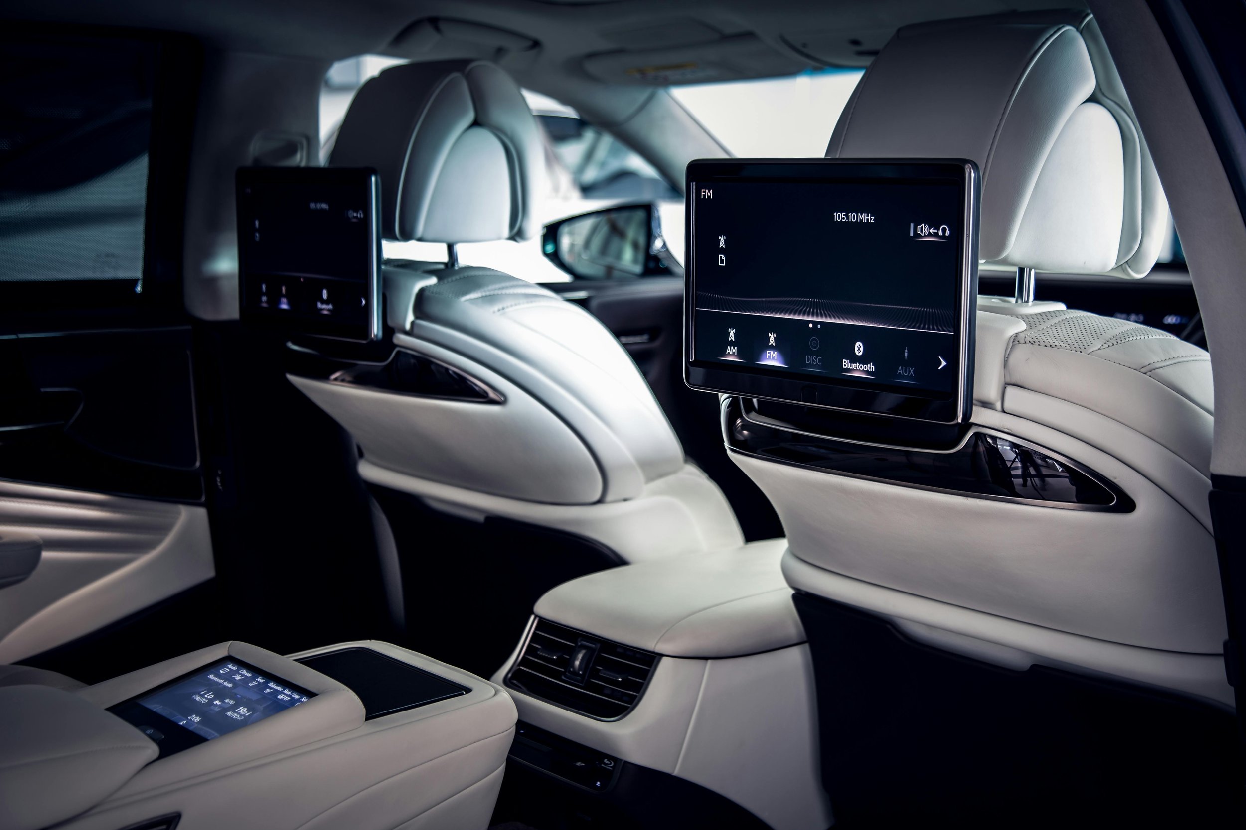 Interior of a luxury car showing the back of the front seats with built-in screens displaying music options, and the beige leather seats and dashboard.