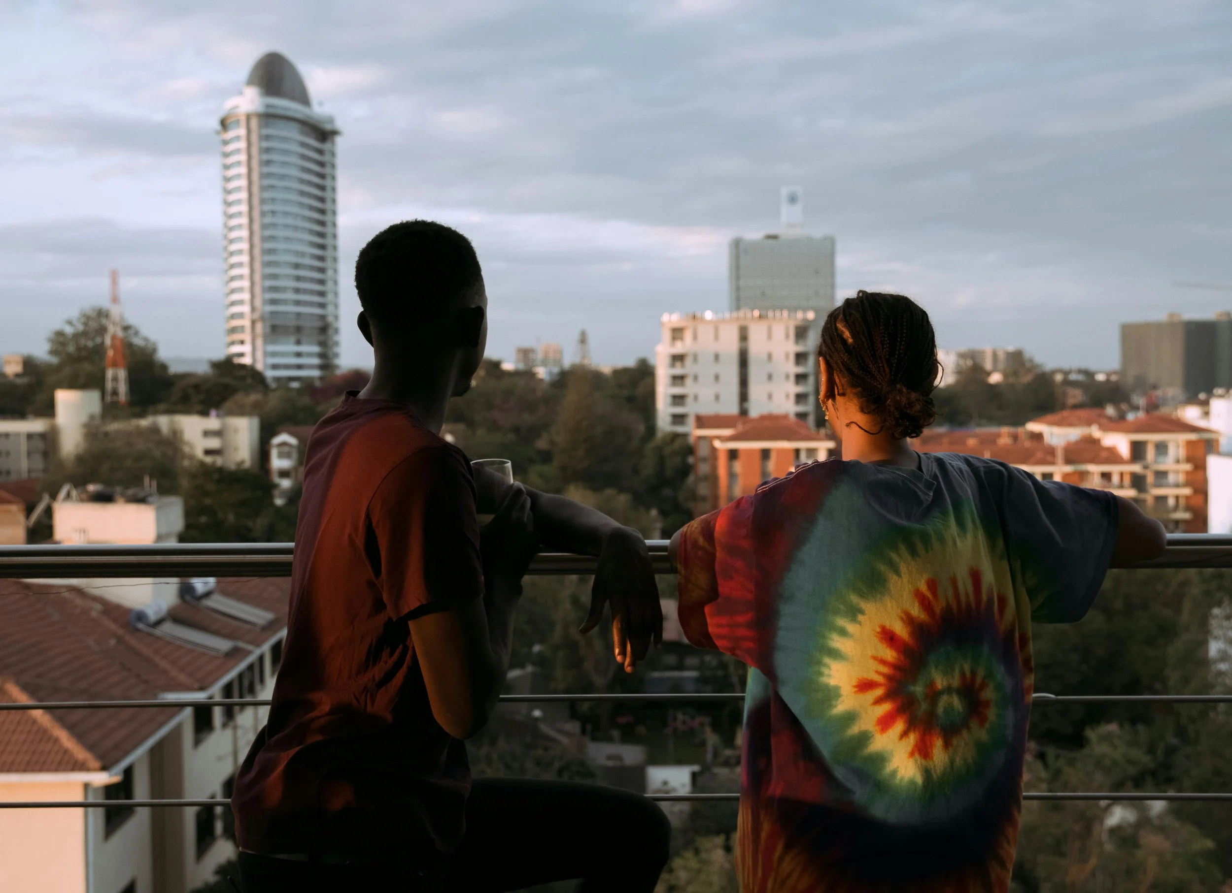 Two people standing by a balcony railing overlooking a cityscape with tall buildings and trees, at sunset or dusk.
