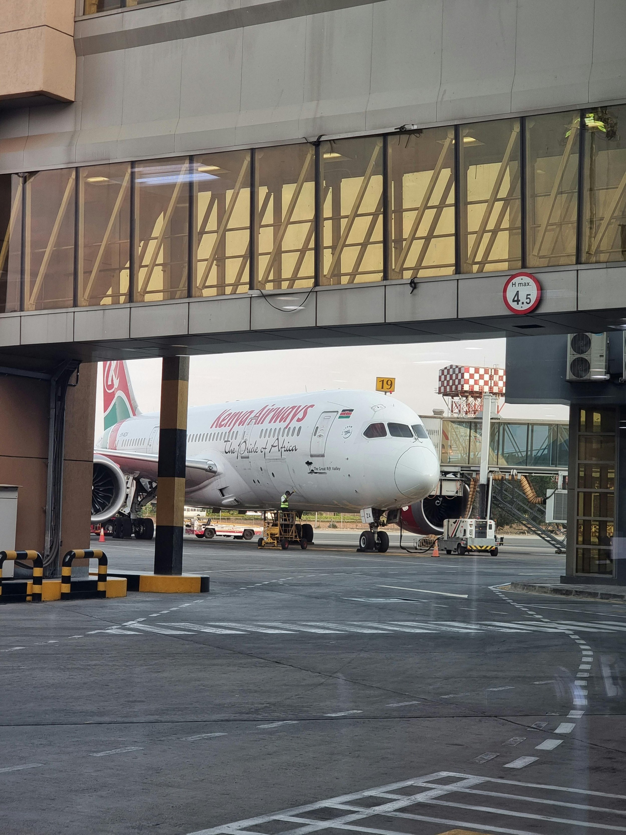 A Kenya Airways airplane parked at the gate with a jet bridge connected. The plane's nose is visible, and ground crew are preparing around it.