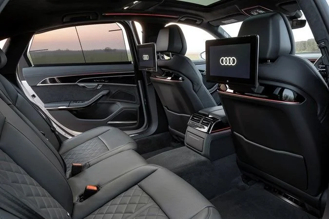 Interior of an Audi vehicle showing black leather seats and two rear-seat entertainment screens mounted on the back of the front seats, with the Audi logo displayed on the screens.