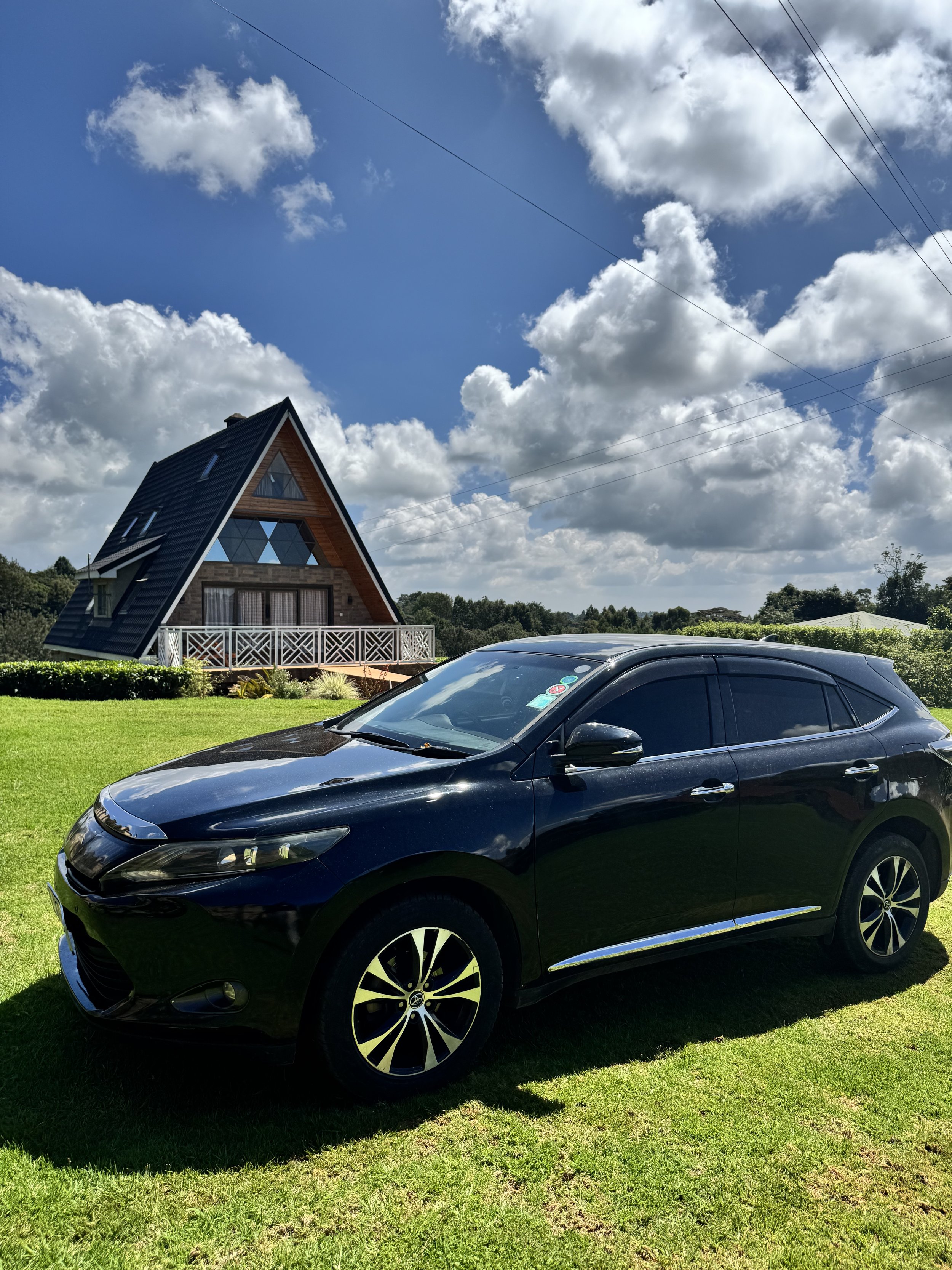 A black crossover SUV parked on a grassy lawn in front of a house with a triangular roof and large windows under a partly cloudy blue sky.