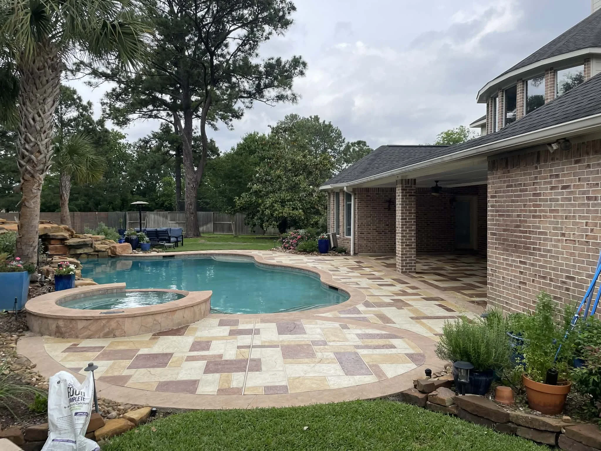 Residential backyard with a swimming pool, hot tub, and patio surrounded by trees, plants, and outdoor furniture.