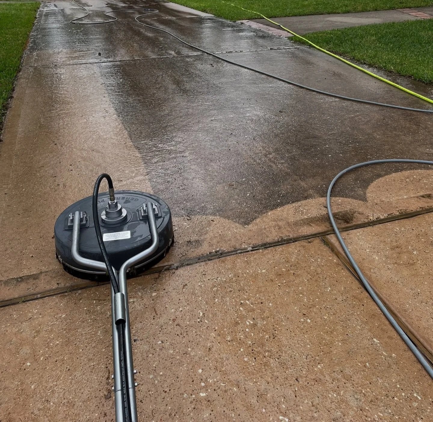A pressure washer cleaning a concrete sidewalk and driveway during rain with hoses connected to the machine.