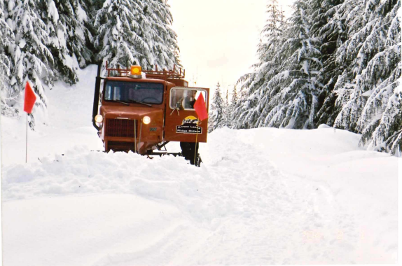 A snowplow working on a snowy forest road, grooming trails with snow-covered trees lining both sides and flag markers along the road, making snowmobiling more accessible.