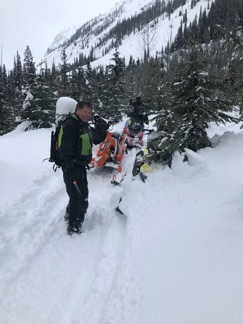 Two people in winter gear with snowmobiles in a snowy mountain forest.